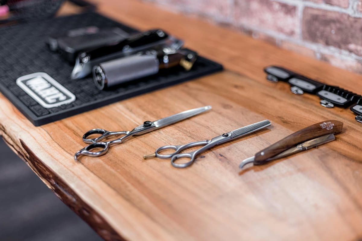 Two pairs of scissors and a wooden-handled straight razor laid on a timber counter beside clippers