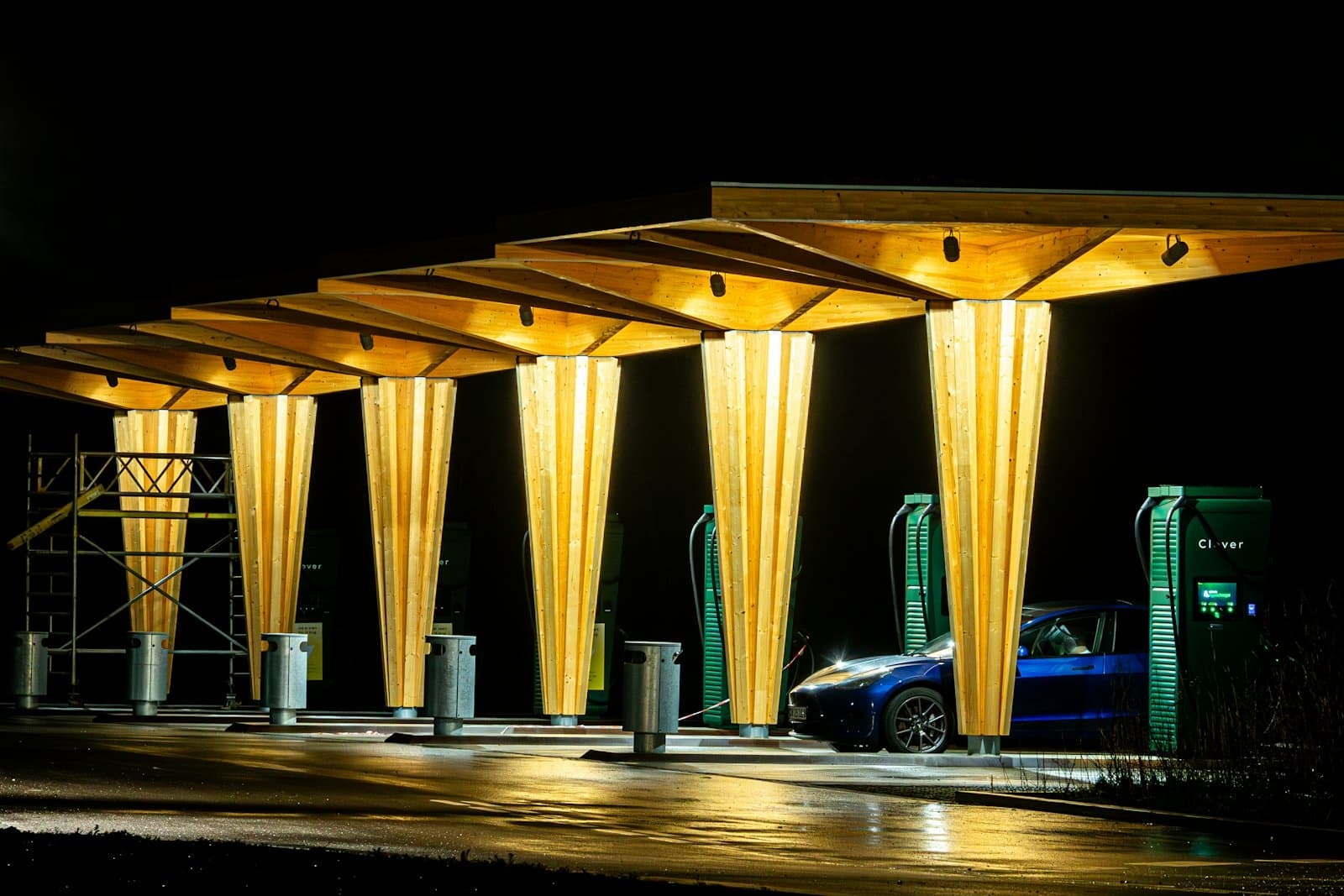 An electric car charging at night under a row of lit timber canopies