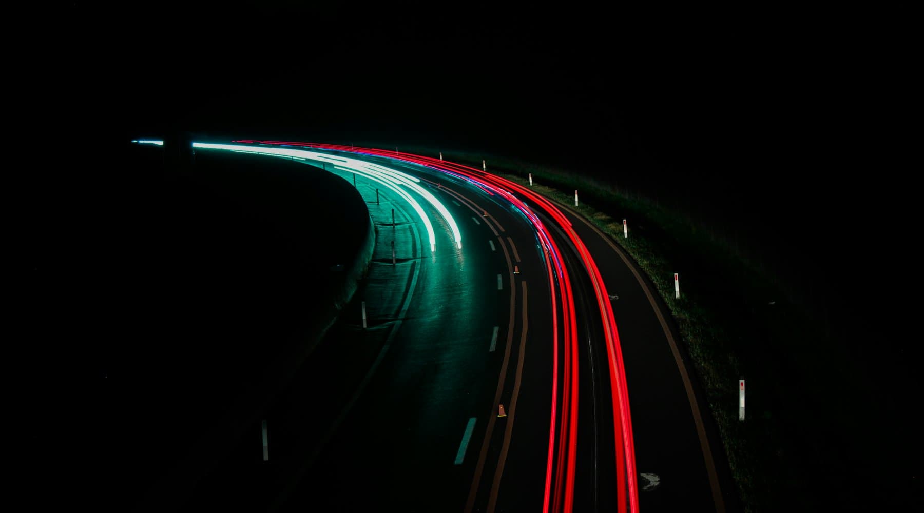 Long-exposure light trails, teal and red, sweeping around a dark bend in the road