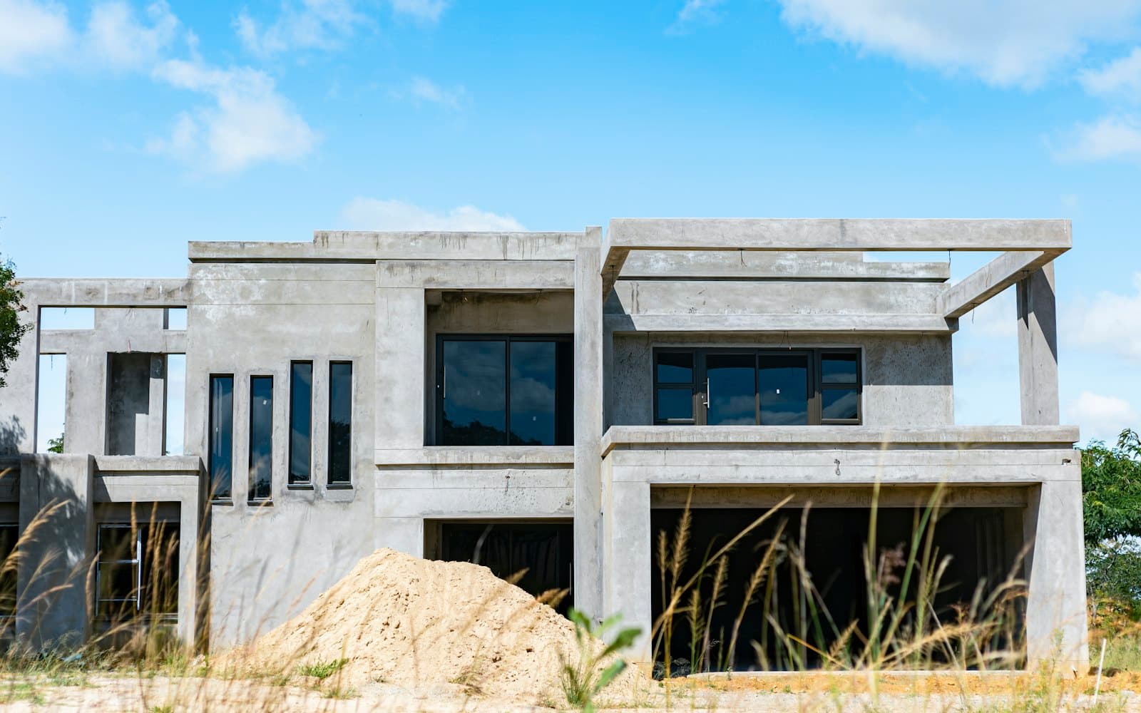 A two-storey concrete house during construction: bare grey frame, windows fitted, a pile of sand in front