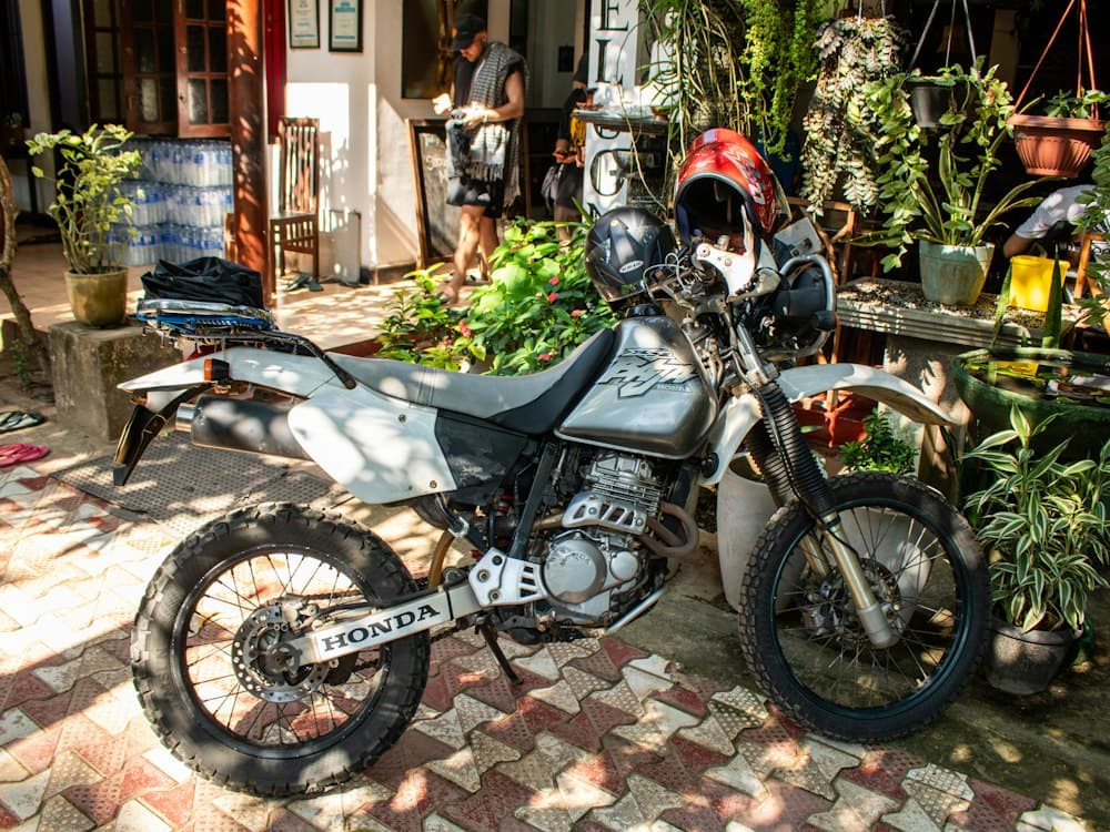 A silver Honda trail bike with knobbly tyres and two helmets on the bars, parked on patterned tiles among potted plants