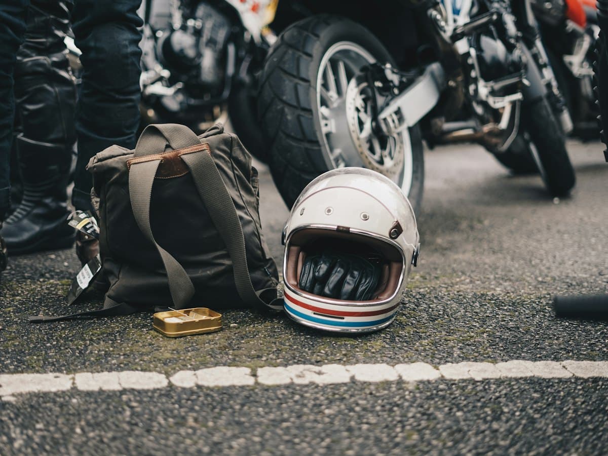 A white open-face helmet with red and blue stripes resting on tarmac beside a canvas rucksack, with parked motorbikes behind