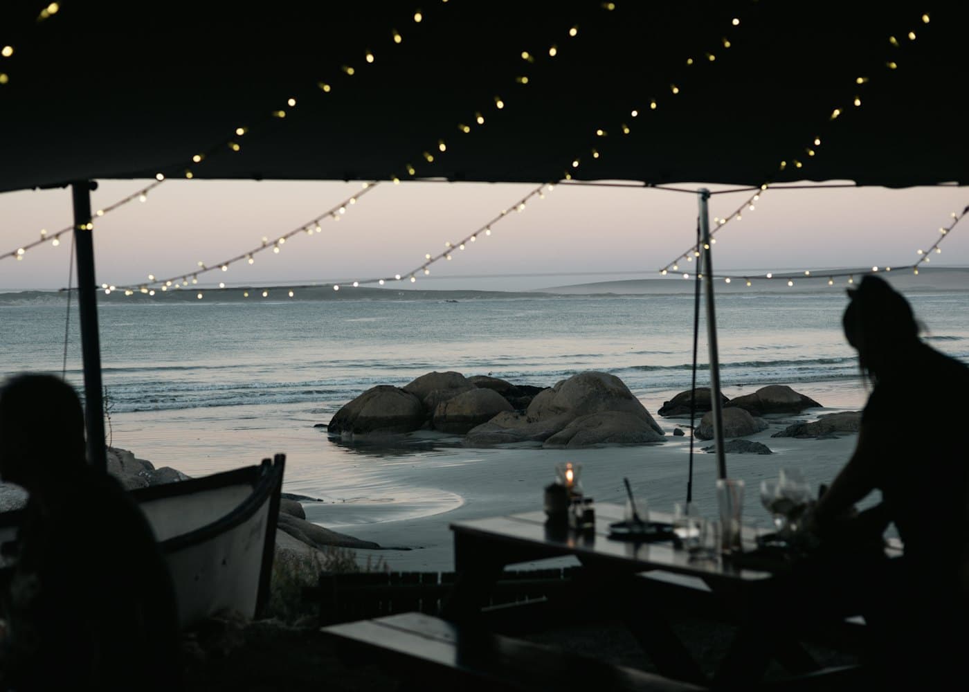 Silhouettes at a candlelit table under strings of small lights, the sea pale behind them