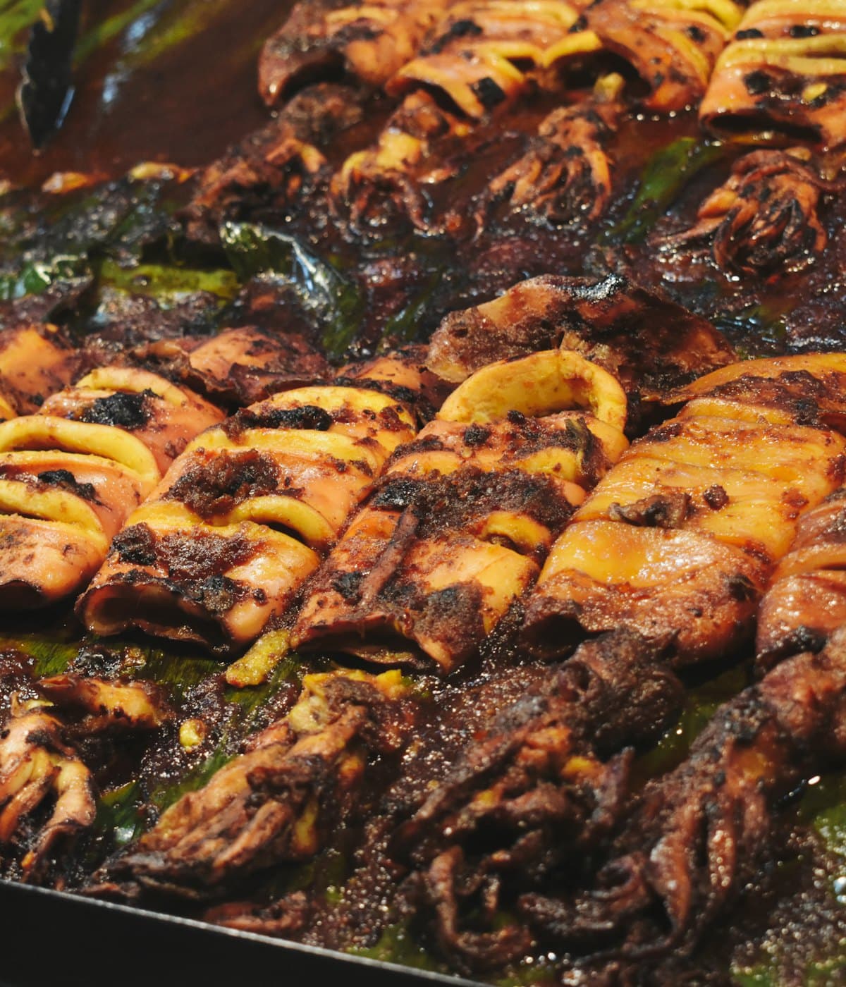 Rows of scored squid on the grill, glazed dark with chilli paste over banana leaf