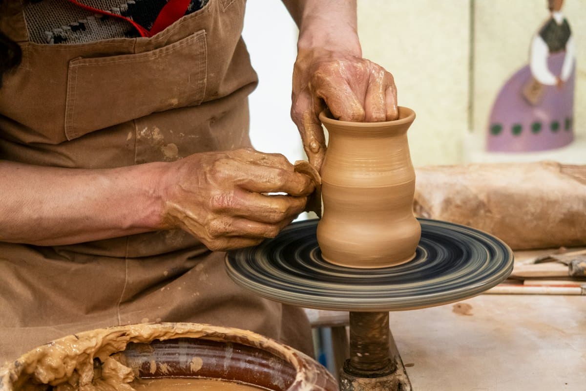 A potter in a clay-stained apron shaping a small pot on a spinning wheel with wet hands