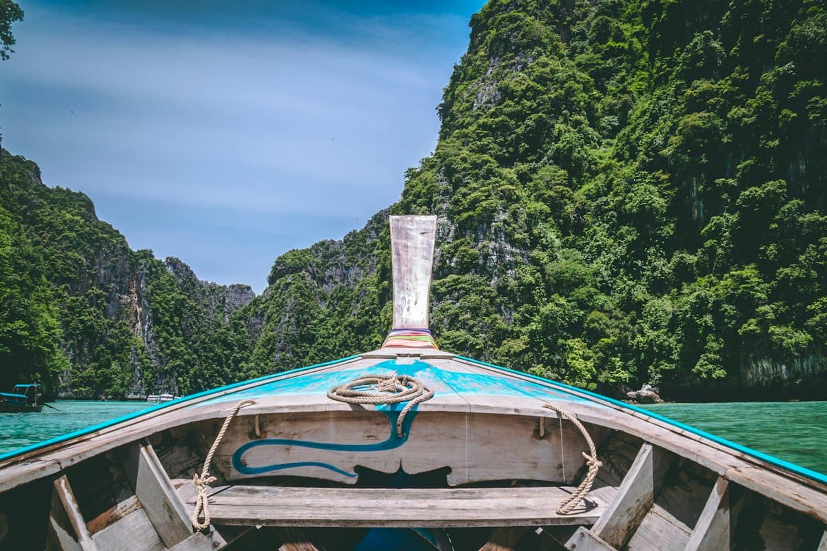 View from the bench of a wooden longtail boat, over its painted bow, towards jungle-covered cliffs