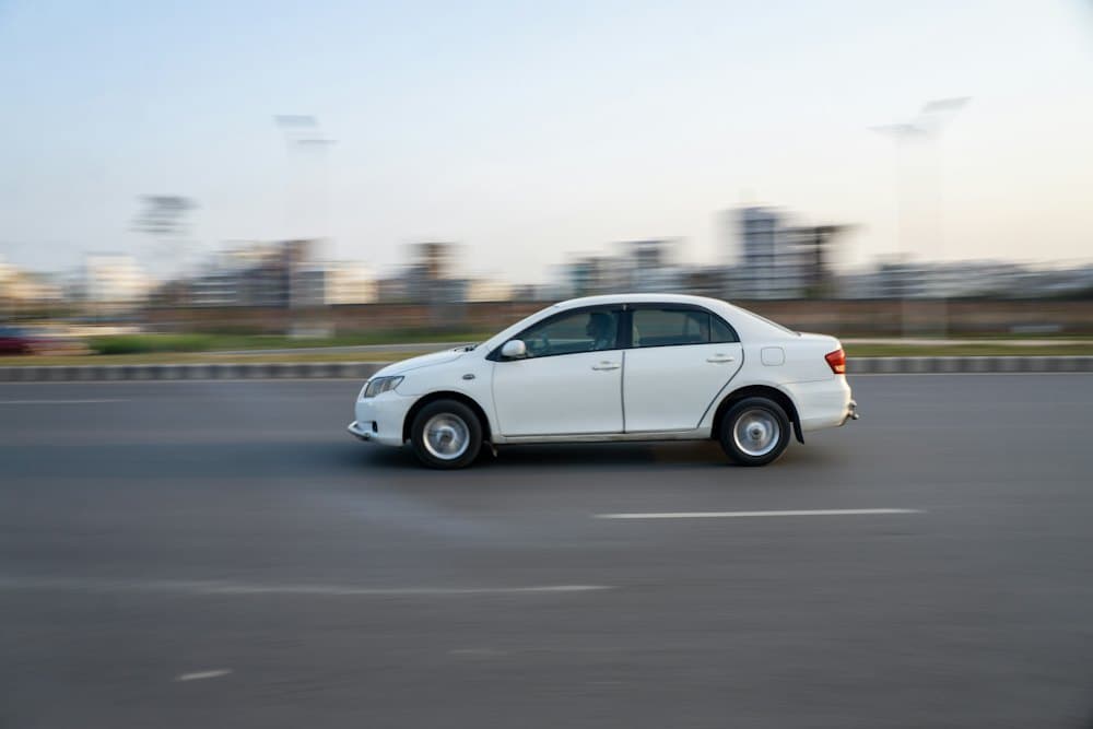 White sedan driving along a wide road at dusk